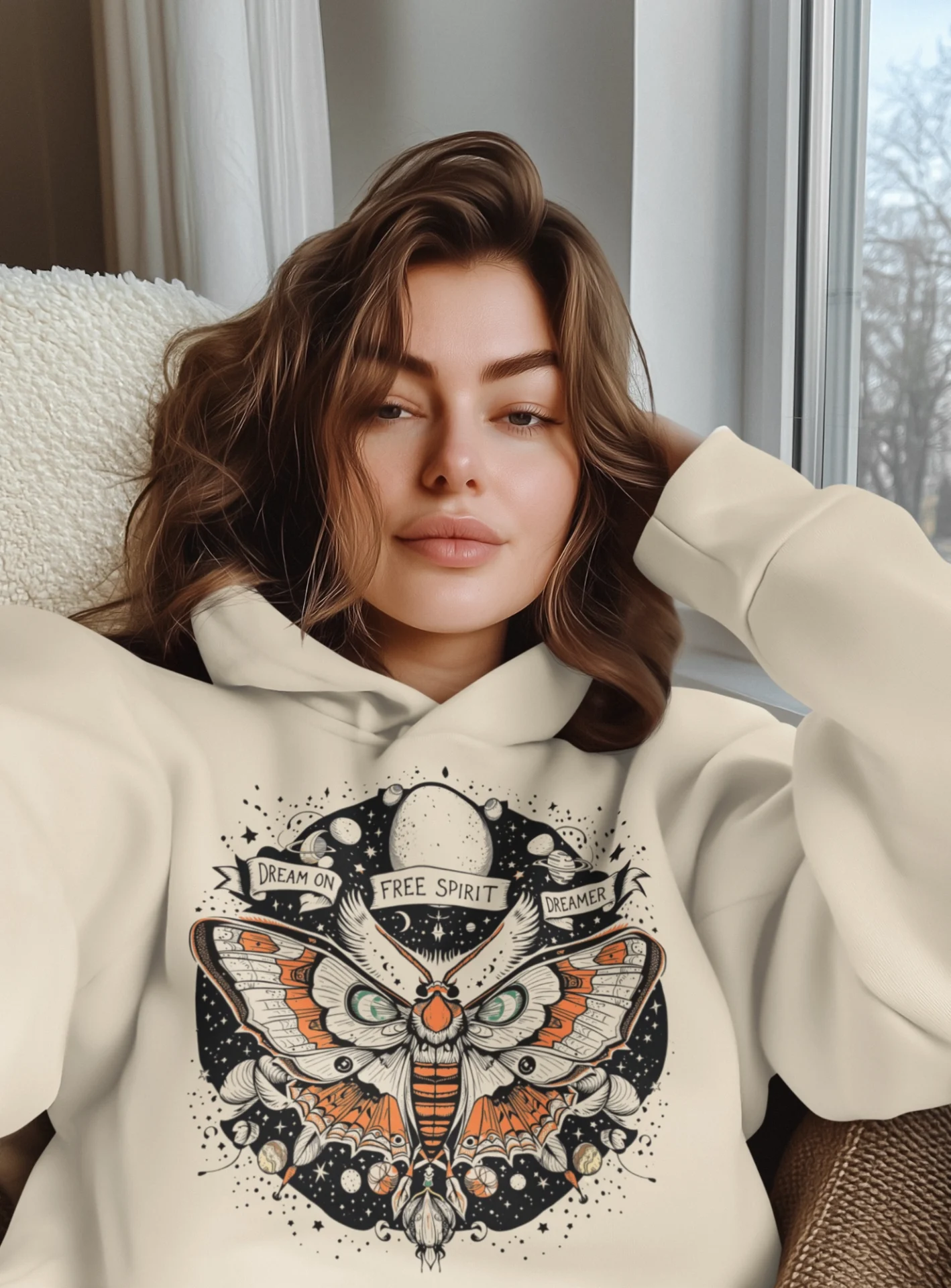 A woman in a beige hoodie with a moth design, relaxed indoors.